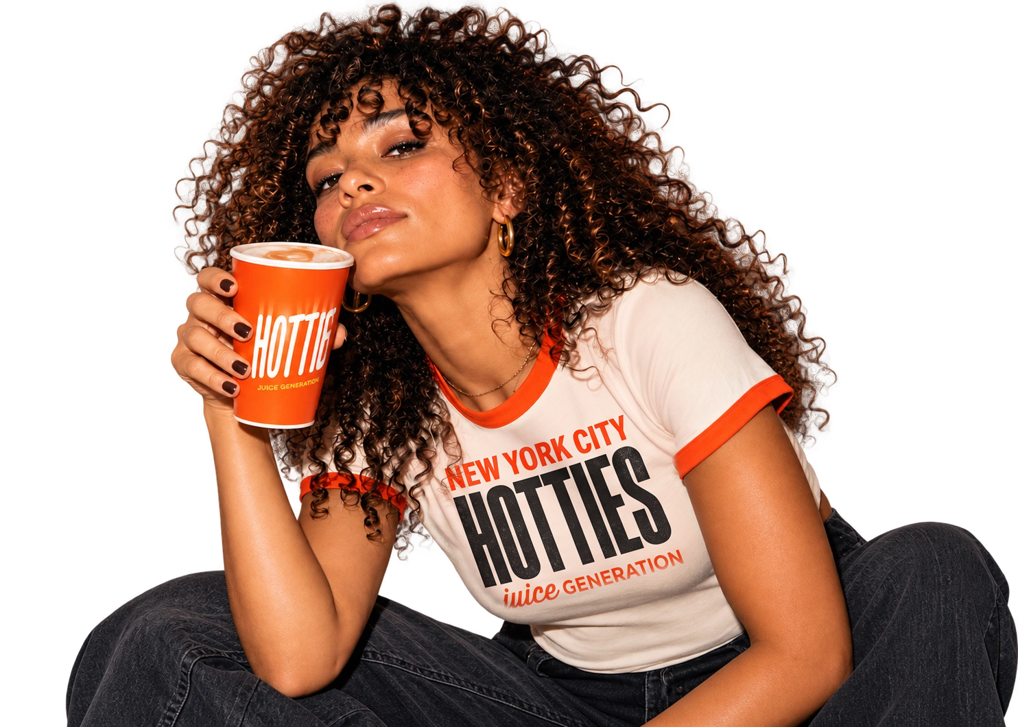 Curly-haired woman wearing the cream HOTTIES ringer tee printed with NEW YORK CITY above HOTTIES and juice GENERATION below, holding an orange HOTTIES cup